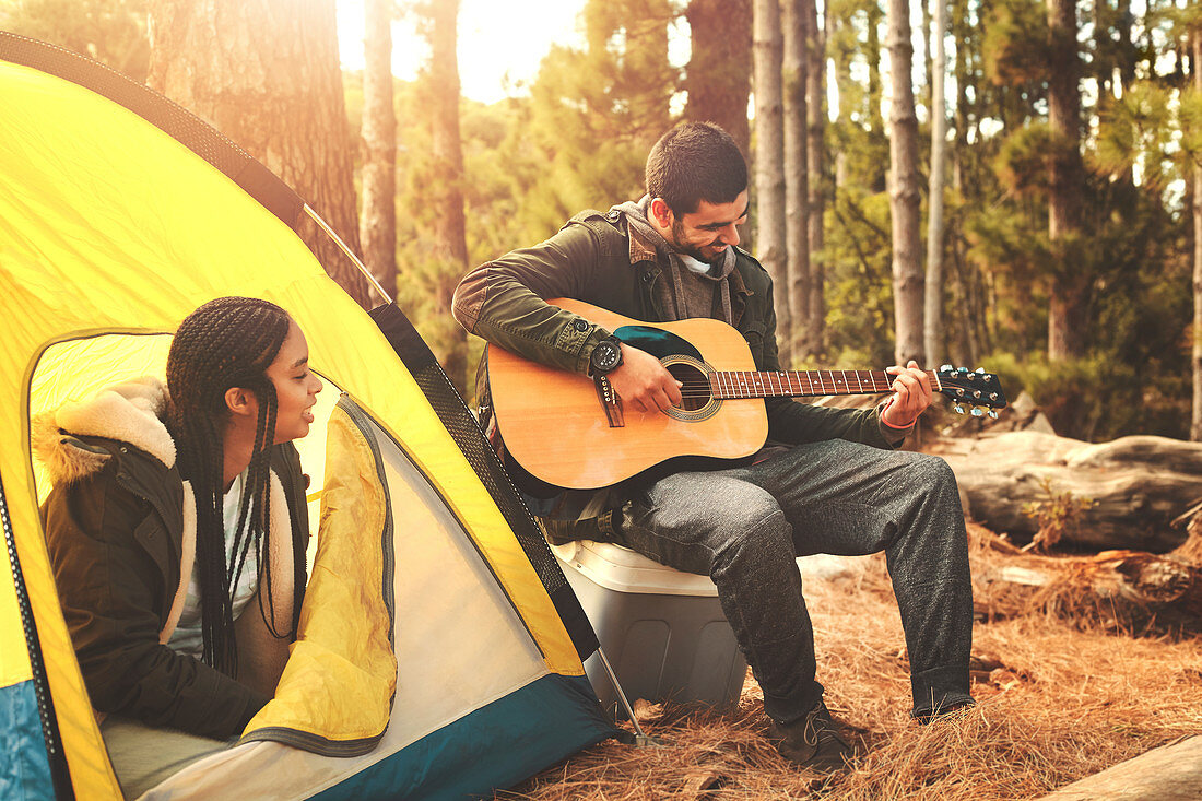 Young couple playing guitar at tent at campsite in woods