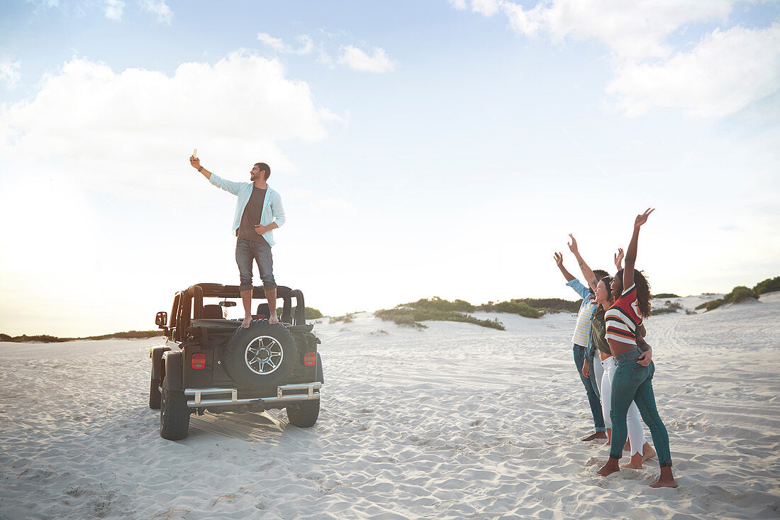 Young man on jeep tire taking selfie with friends on beach