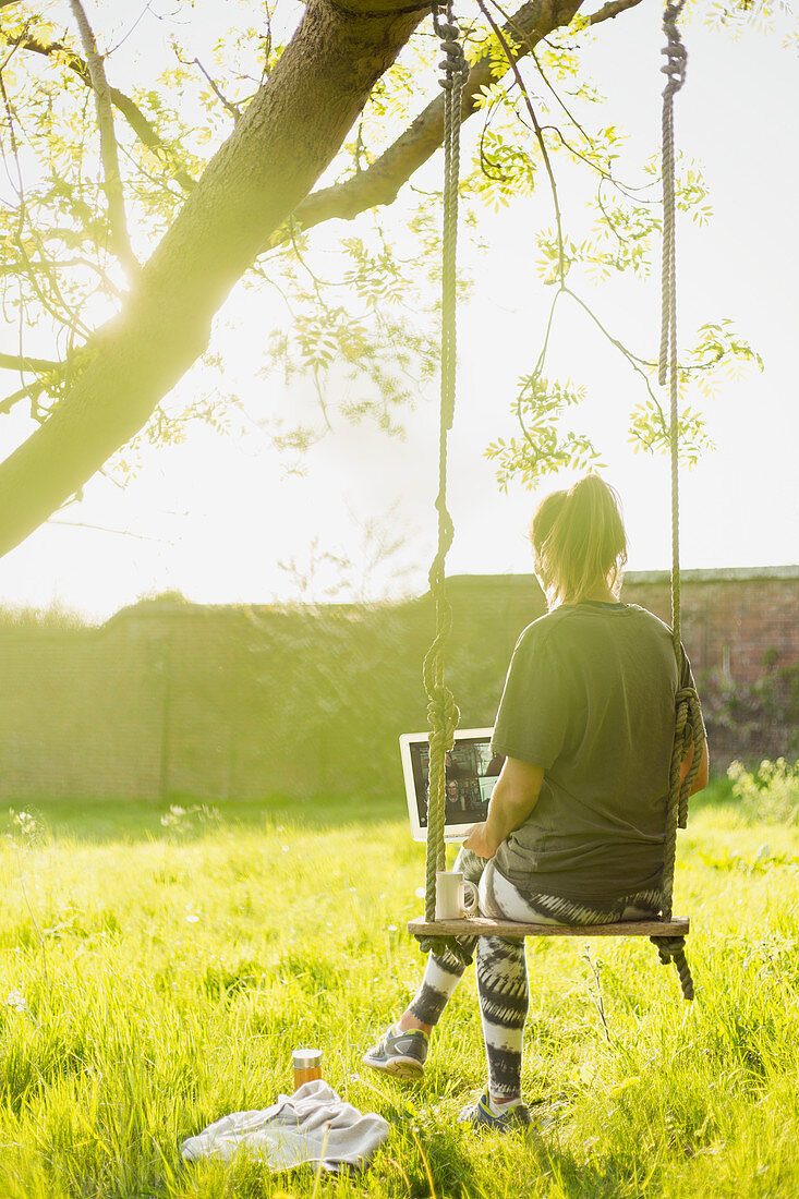 Woman with laptop video chatting on swing in sunny garden