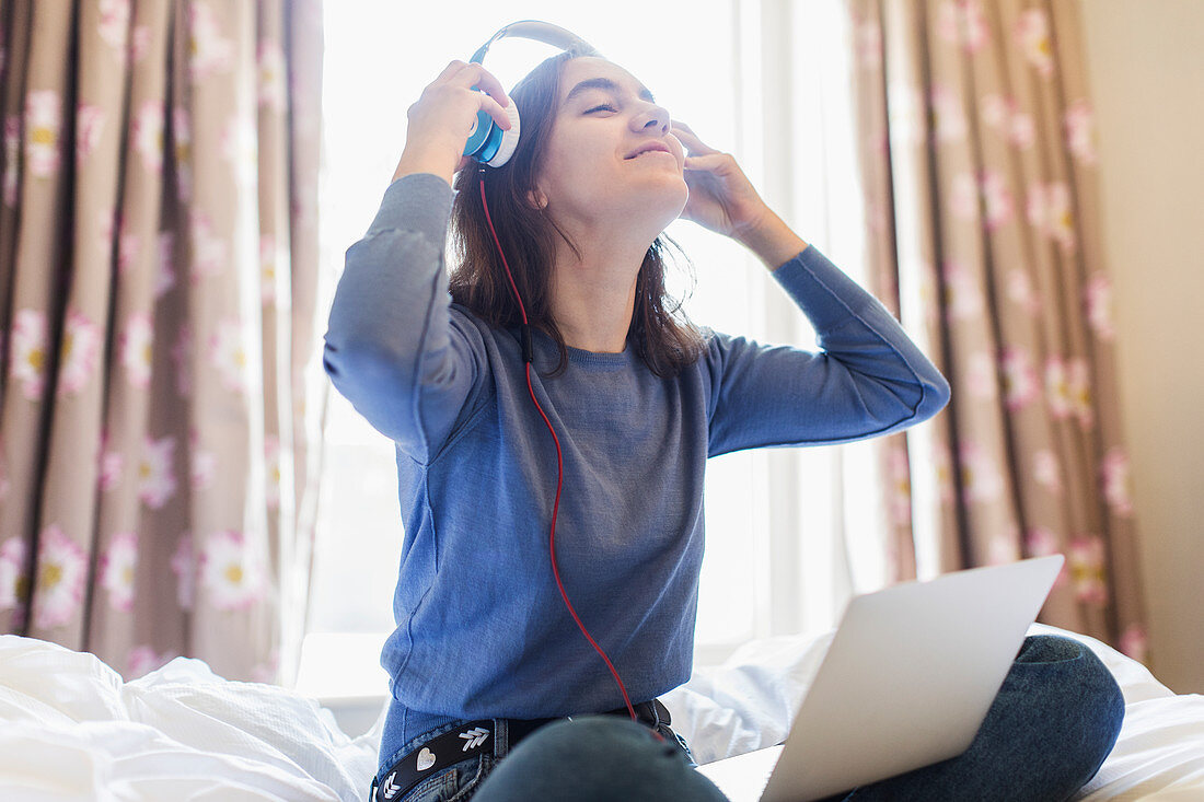 Smiling teenage girl with headphones and laptop on sunny bed