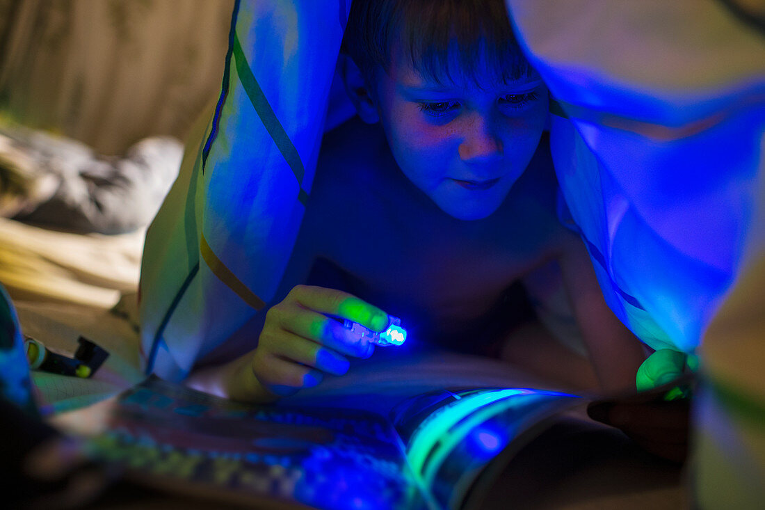 Boy with black light reading magazine under blanket