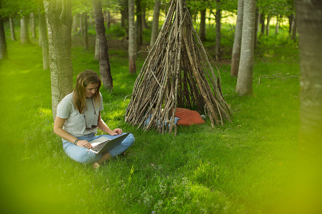 Young woman using laptop at branch teepee in woods