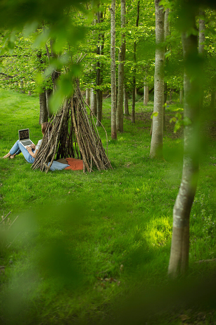 Woman using laptop at branch teepee in woodland