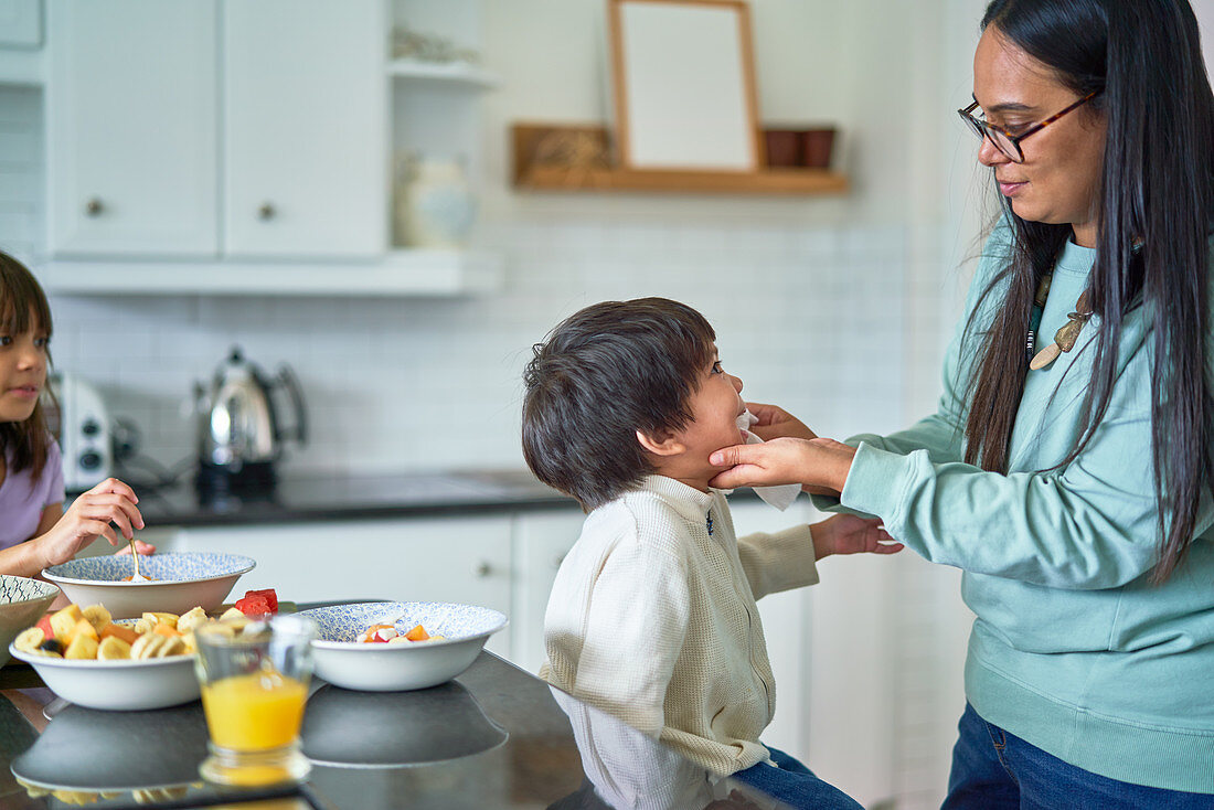 Mother wiping face of son eating fruit in kitchen