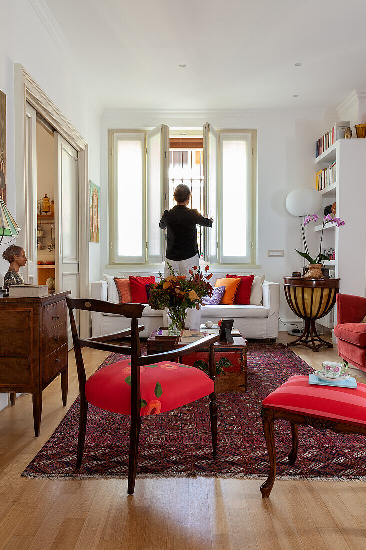 Red chairs, antique chest of drawers, chests used as coffee table and sofa in living room; woman in background