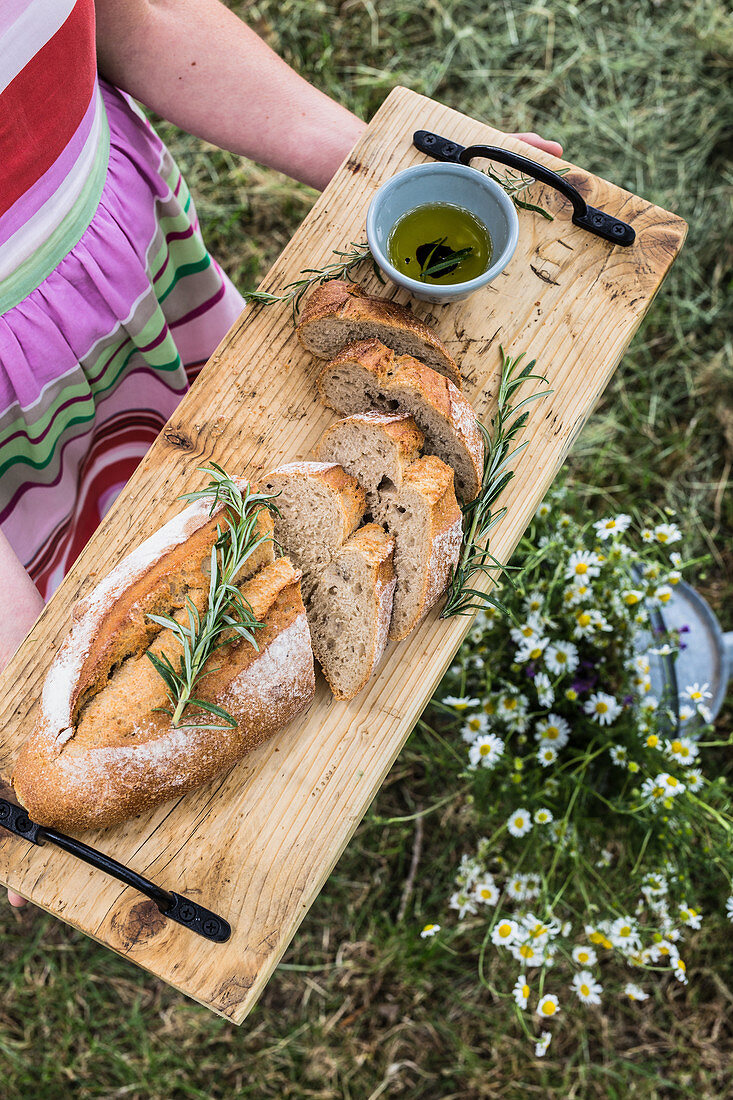 Female holding wooden cutting board with bread cut on loafs with fresh rosemary and olive oil
