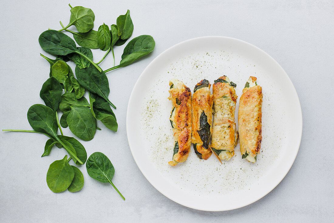 Rolls of roasted chicken fillet placed on plate near fresh spinach leaves on light gray table
