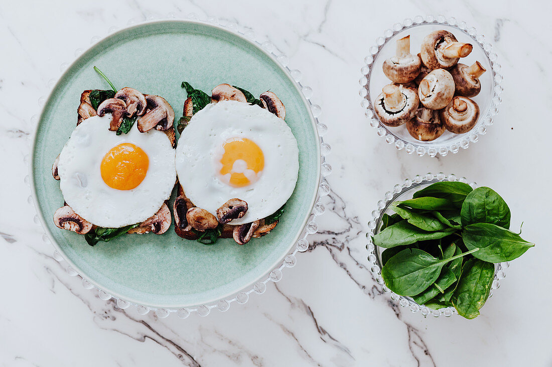 Fried eggs placed of sandwiches with mushrooms and spinach on plate on marble table