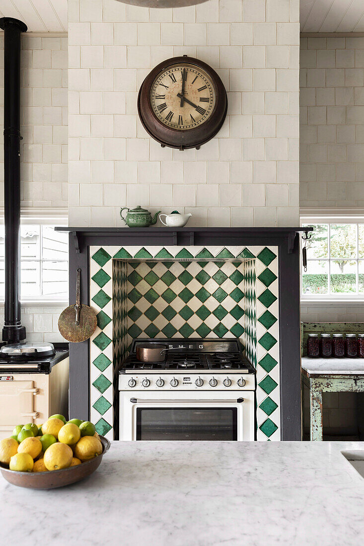 View over kitchen island with marble worktop to gas cooker in wall niche with green-white tiles, above kitchen clock