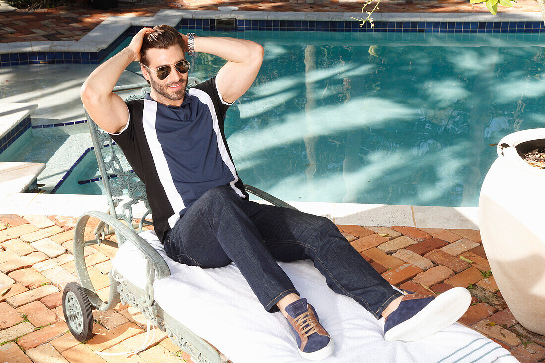 Young man with a beard in a polo shirt and jeans is sitting on a lounger by the pool