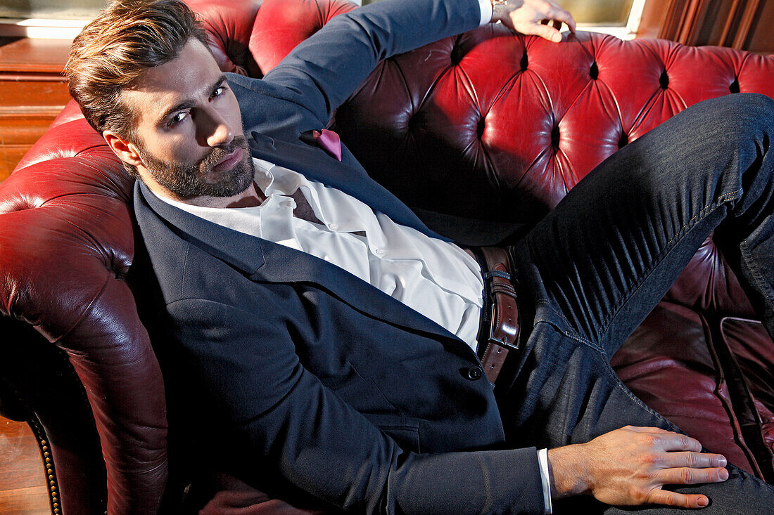 Young man with a beard in a jacket and jeans lies on a leather sofa