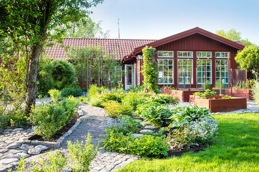 Frühlingsgarten mit Pflastersteinen und Kiesweg, im Hintergrund rot gestrichenes Holzhaus