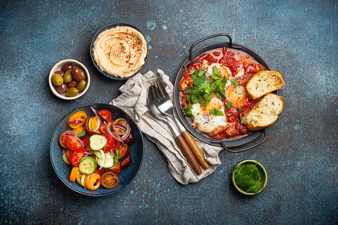 Shakshouka in der Pfanne mit Röstbrot, Gemüsesalat, Hummus und Oliven (traditionelles Frühstück oder Brunch aus dem Nahen Osten)