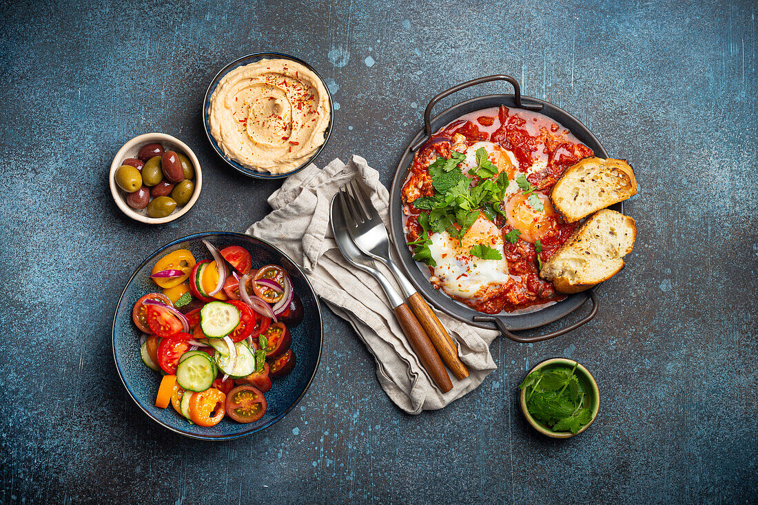 Shakshouka in pan with toasts, fresh vegetables salad, hummus and olives (Middle Eastern traditional breakfast or brunch)