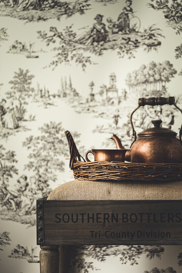 Tea kettle and cup on wicker tray in front of Toile de jouy wallpaper