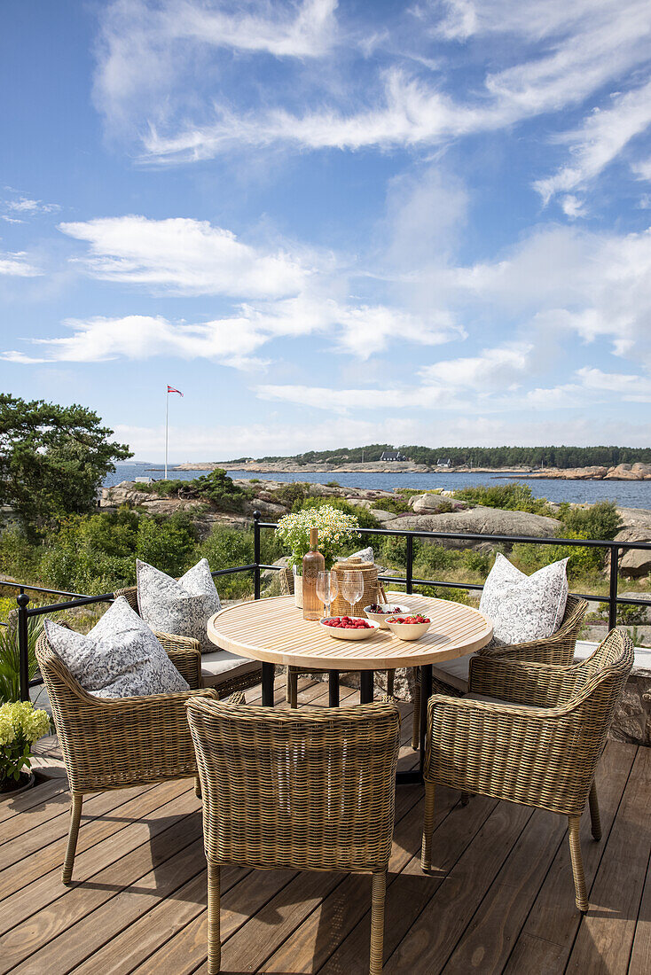 Round table with rattan chairs and cushions on terrace, view of coastal landscape