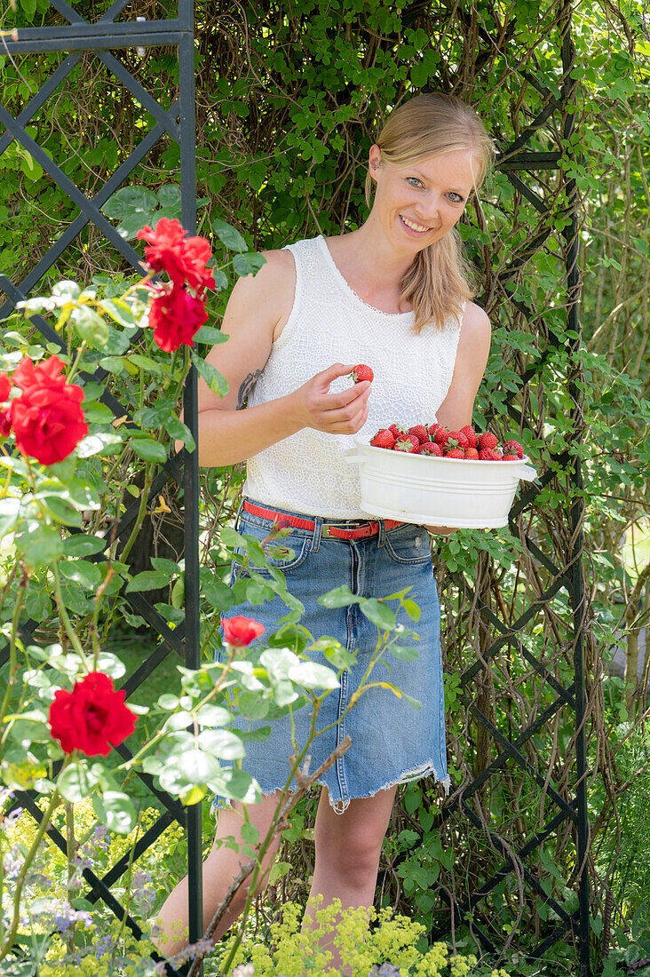 Frau mit frischen Erdbeeren in Schale unter Rosenbogen im sommerlichen Garten