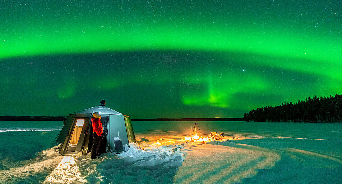 Wanderer beobachtet die Aurora Borealis (Nordlichter) in der Nähe von Lagerfeuer und Iglu in der gefrorenen Landschaft, Jokkmokk, Lappland, Schweden, Skandinavien, Europa