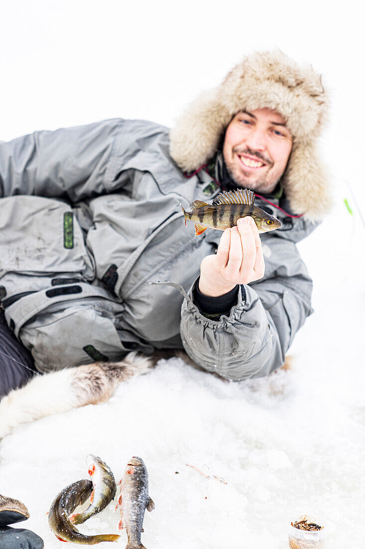 Porträt eines lächelnden Mannes, der einen nach dem Eisfischen gefangenen Fisch hält, Lappland, Schweden, Skandinavien, Europa