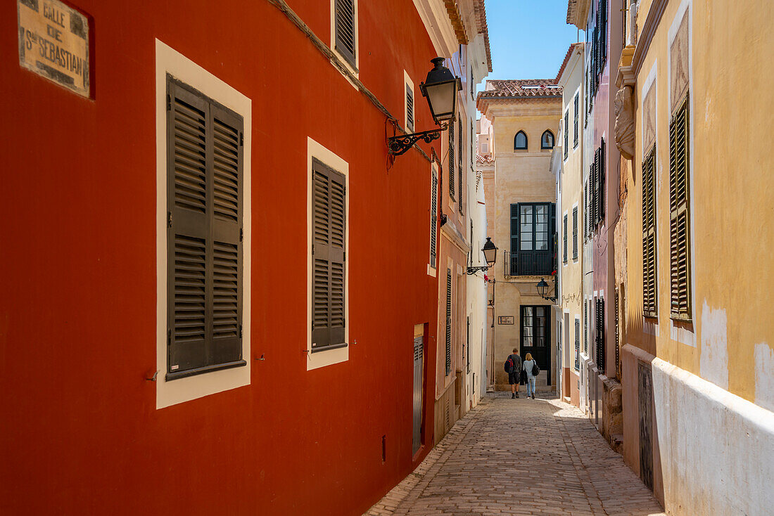 Blick auf ein Paar, das eine pastellfarbene Straße im historischen Zentrum entlanggeht, Ciutadella, Menorca, Balearen, Spanien, Mittelmeer, Europa