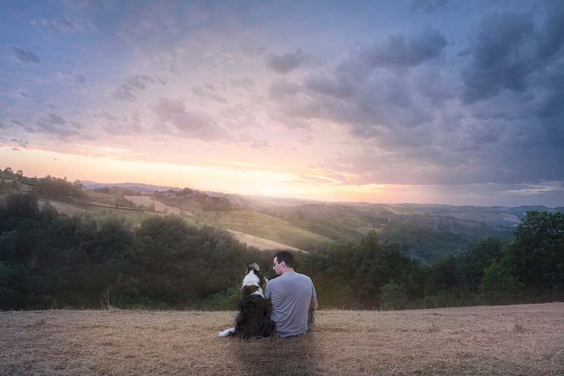 Ein Mann und sein Border Collie Hund sitzen auf einem Feld in der Landschaft und genießen den Sonnenuntergang, Italien, Europa
