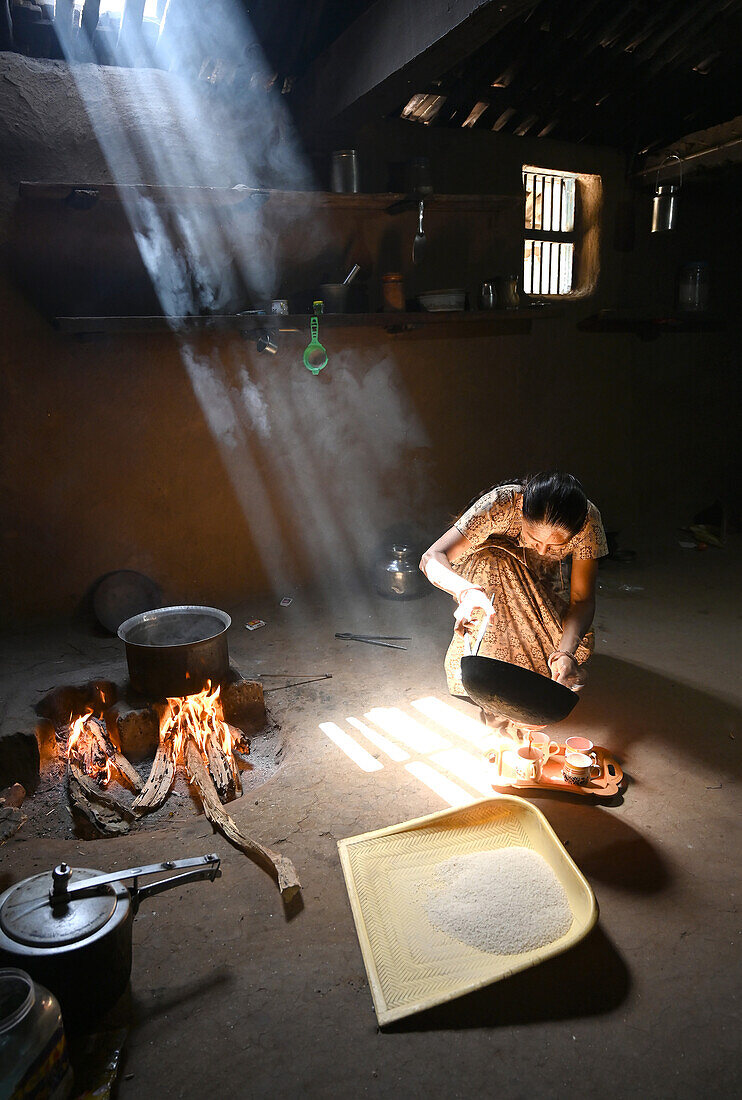Sonnenlicht fällt in eine Küche mit Lehmwänden, Frau macht Chai, Topf auf Holzfeuer, Reisstreuer auf dem Boden, Kadana, Gujarat, Indien, Asien