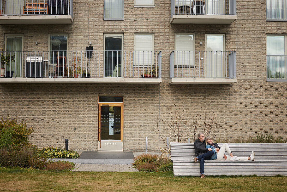 Couple relaxing on bench in front of block of flats
