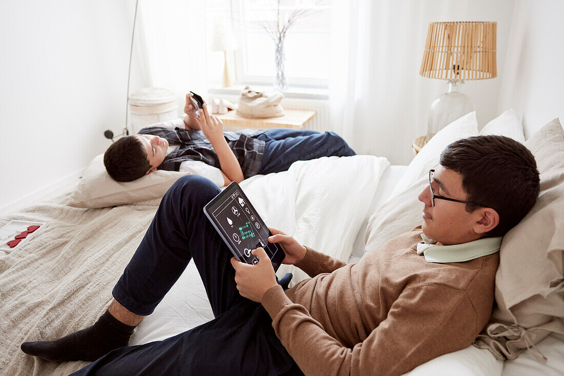 Brothers lying on bed and using phone and tablet