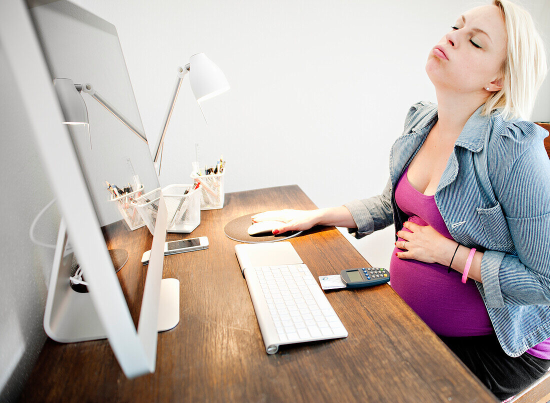 Schwangere Frau sitzt am Schreibtisch und fühlt sich krank
