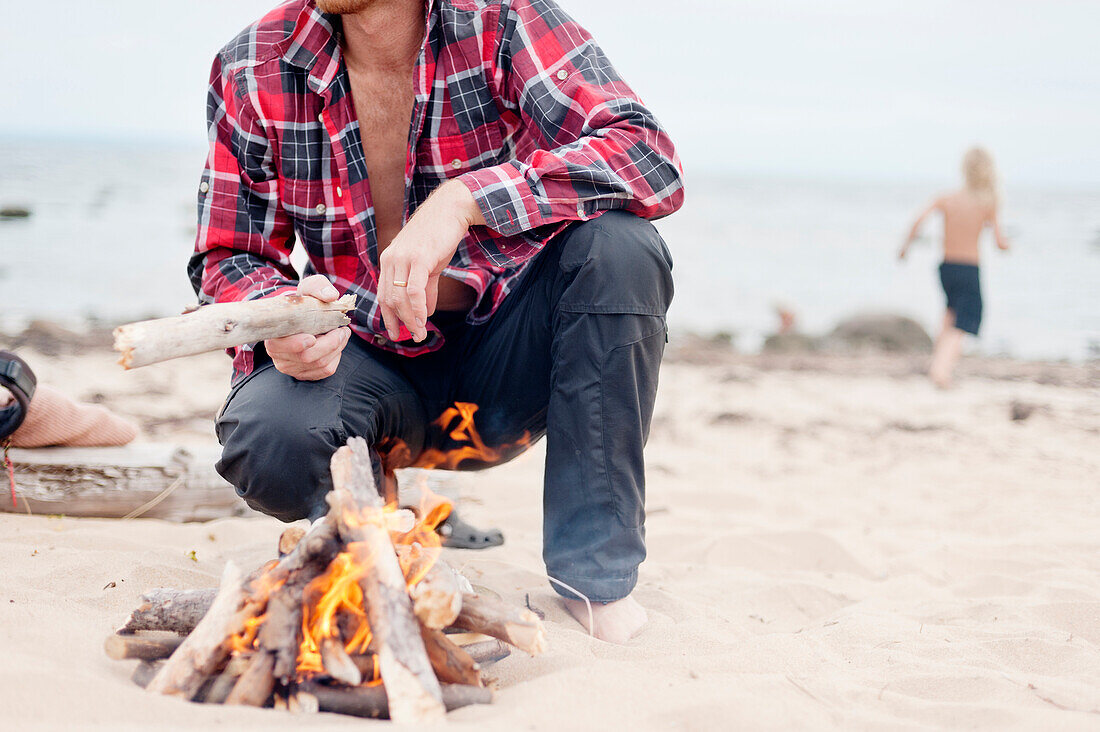 Mann kauert neben einem Lagerfeuer am Strand