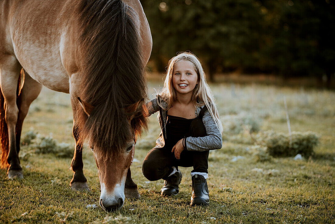 Lächelndes Mädchen hockt auf einer Wiese mit grasendem Pony