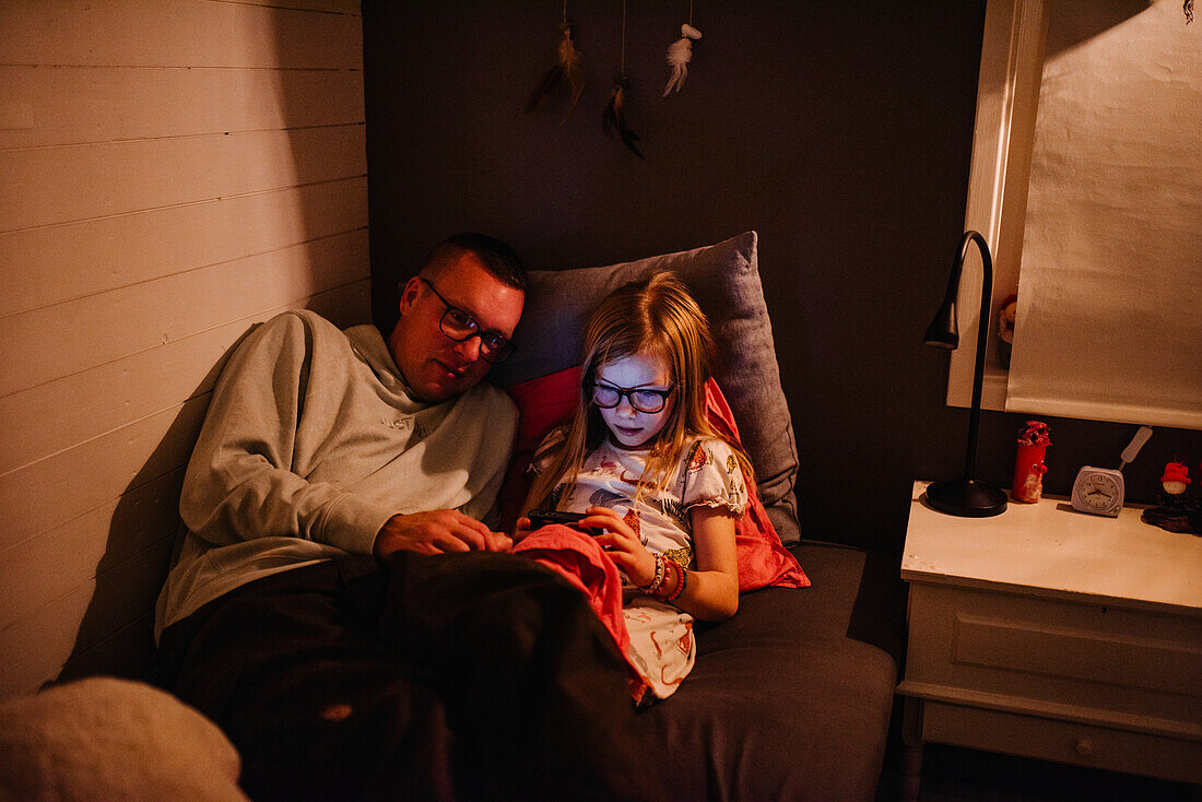 Vater mit Tochter, die auf dem Bett sitzt und auf ihr Handy schaut