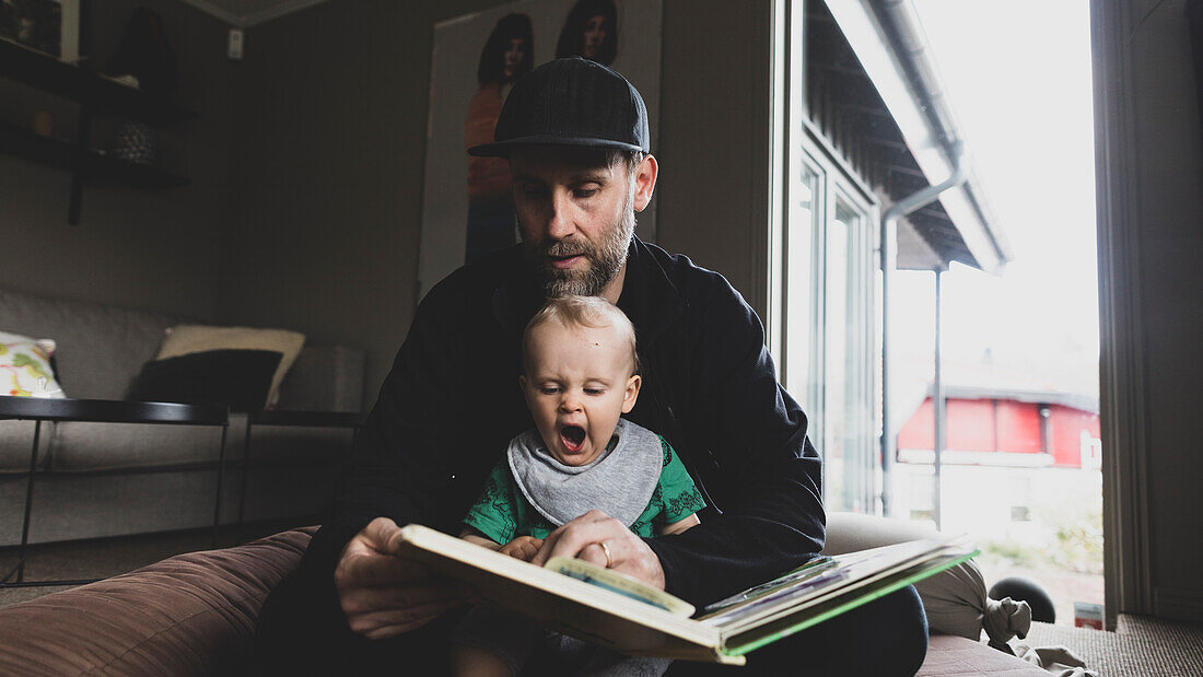Father reading book for yawning baby son