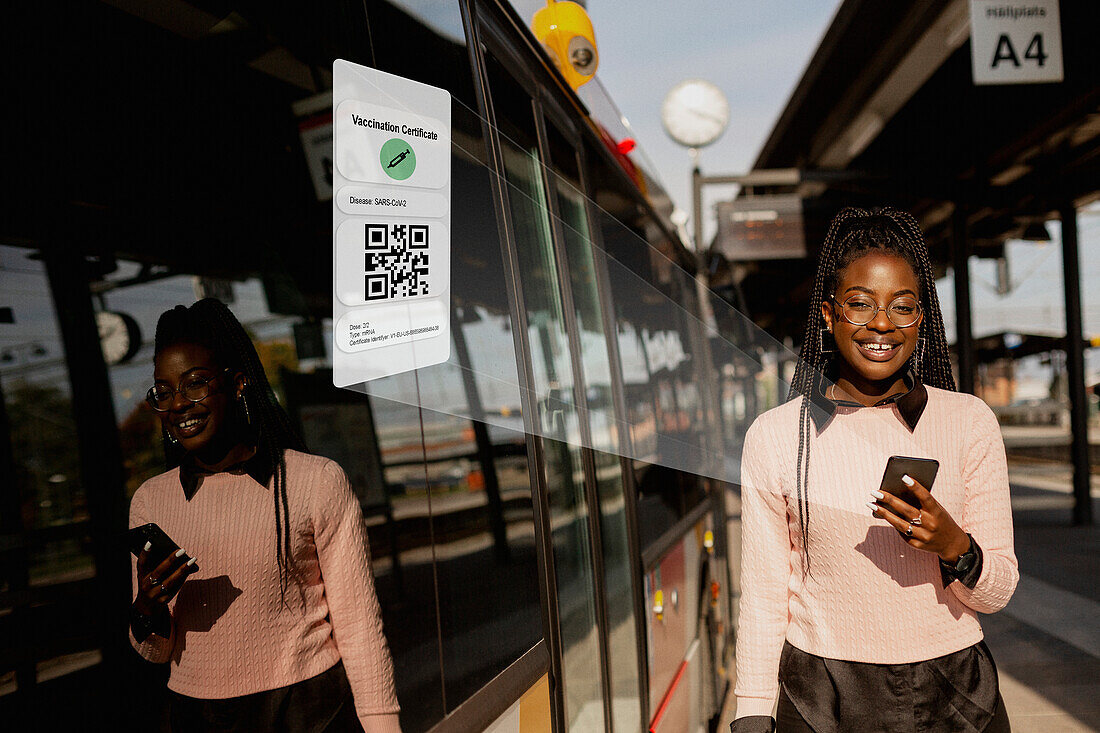 Young woman using cell phone at bus station with Covid-19 vaccine certificate