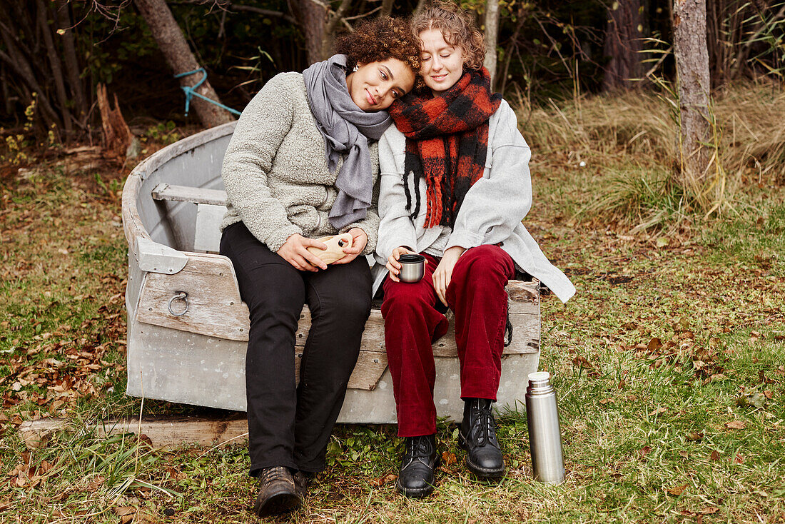 Junges weibliches Paar sitzt auf einem alten Boot und trinkt heiße Getränke