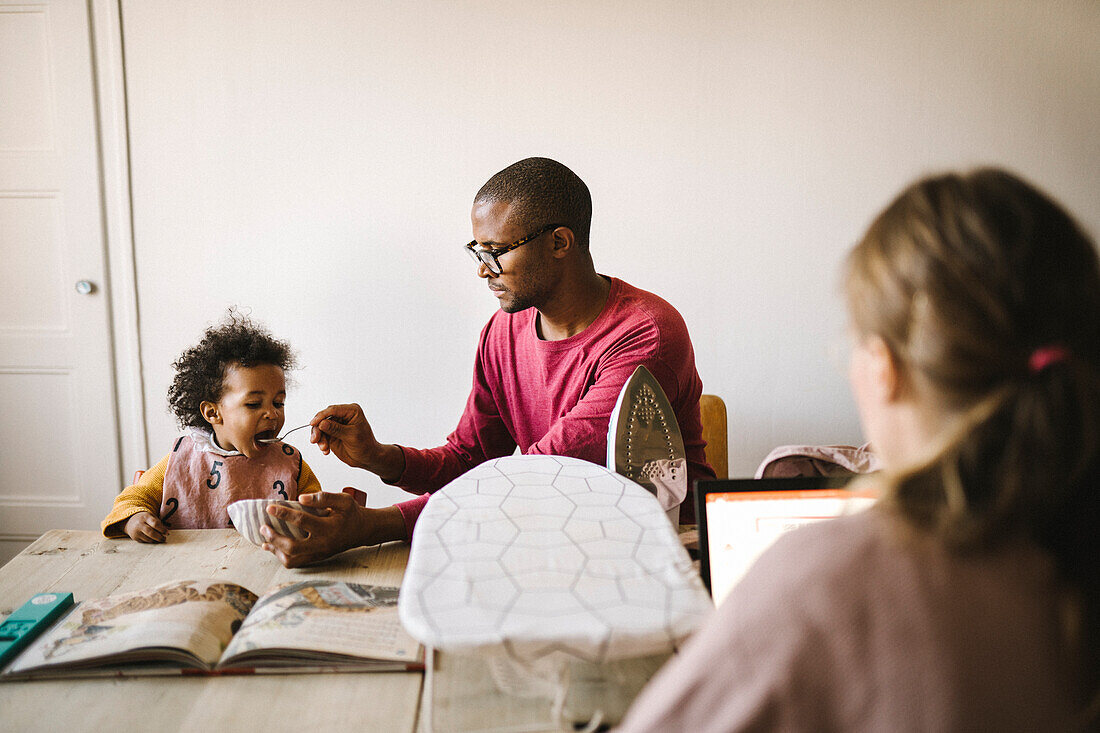 Father feeding daughter while mother working on laptop