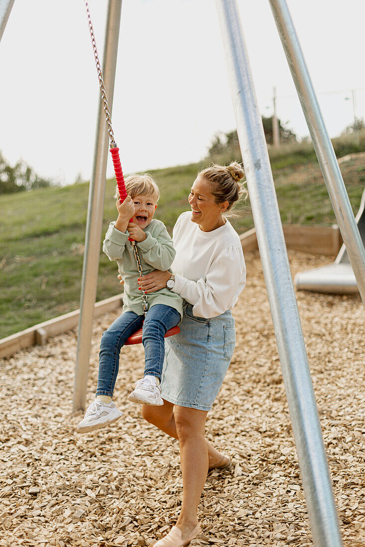 Glückliche Mutter schaukelt ihren Sohn auf dem Spielplatz