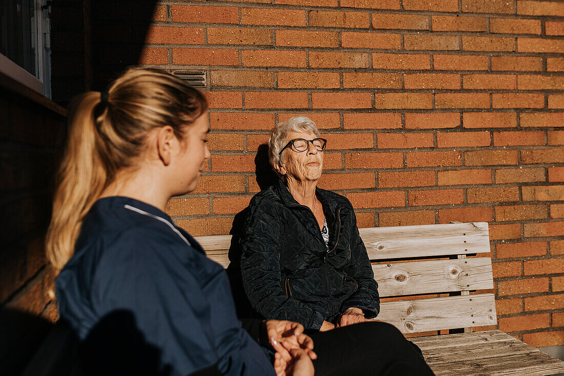 Home caretaker and senior woman sitting on bench and sunbathing