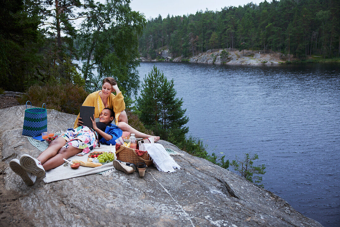 Weibliches Paar picknickt am Fluss und benutzt ein Tablet