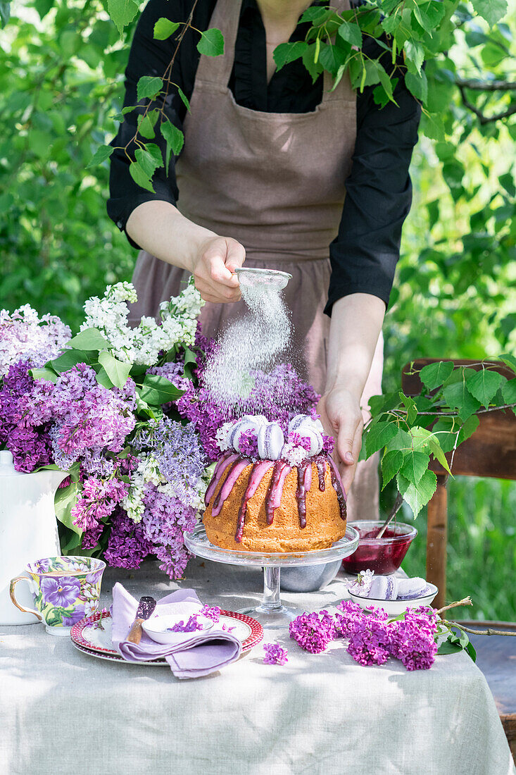 Kuchen mit Heidelbeerglasur und Macarons auf Gartentisch neben Fliederstrauß