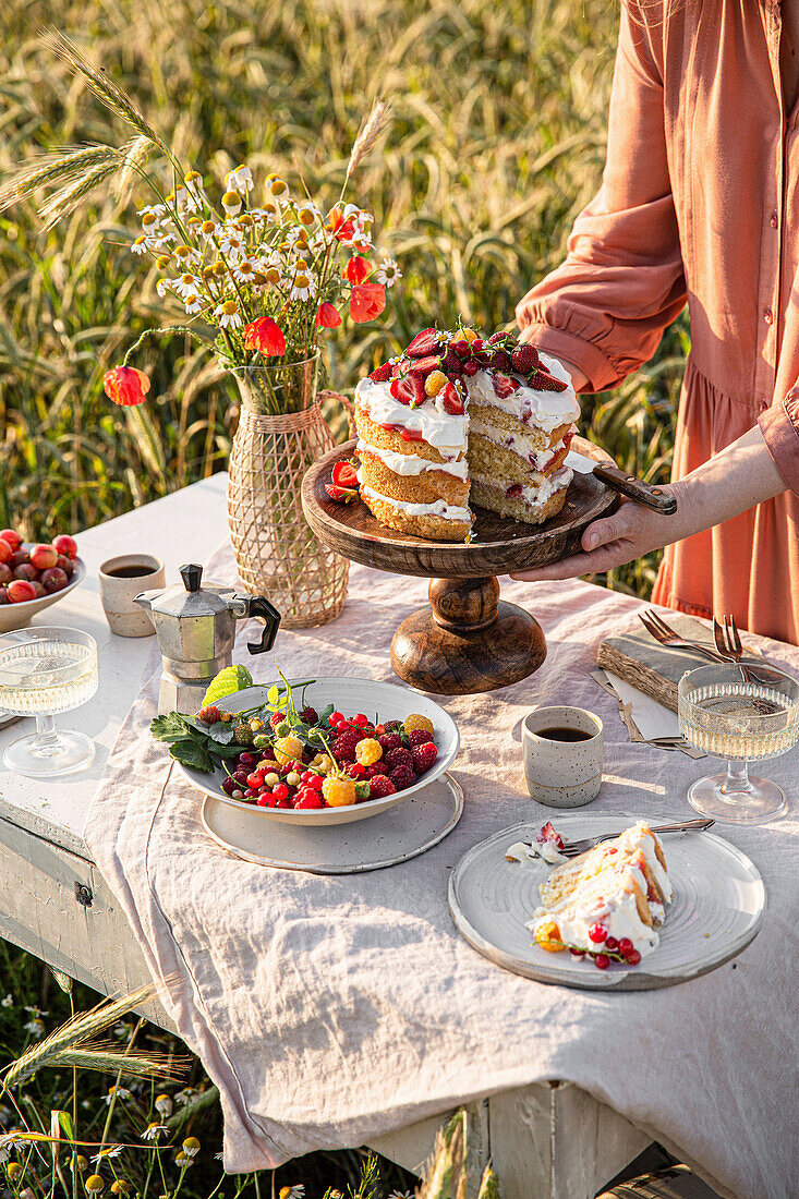 Gedeckter Tisch mit Kuchen, Blumenstrauß und Beeren mitten im Feld