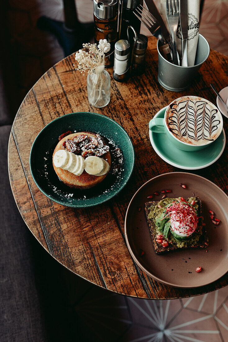 Avocado toasted topped with an egg, pancakes topped with banana and nuts, and a latte on breakfast table