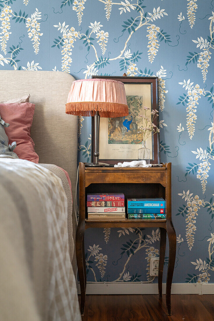 Nachttisch mit Büchern und Lampe vor floraler Tapete