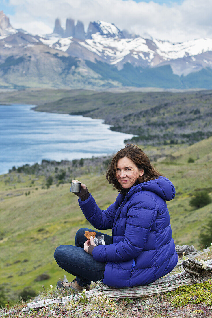 Eine Frau sitzt bei einem Getränk und einem Snack mit Blick auf einen See und eine zerklüftete Berglandschaft, Torres Del Paine Nationalpark; Torres Del Paine, Magallanes und Antartica Chilena Region, Chile