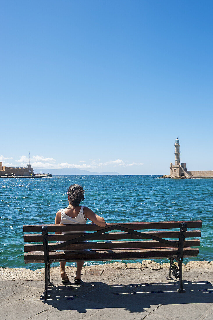 Eine Frau sitzt auf einer Bank mit Blick auf den Hafen und den Leuchtturm; Chania, Kreta, Griechenland