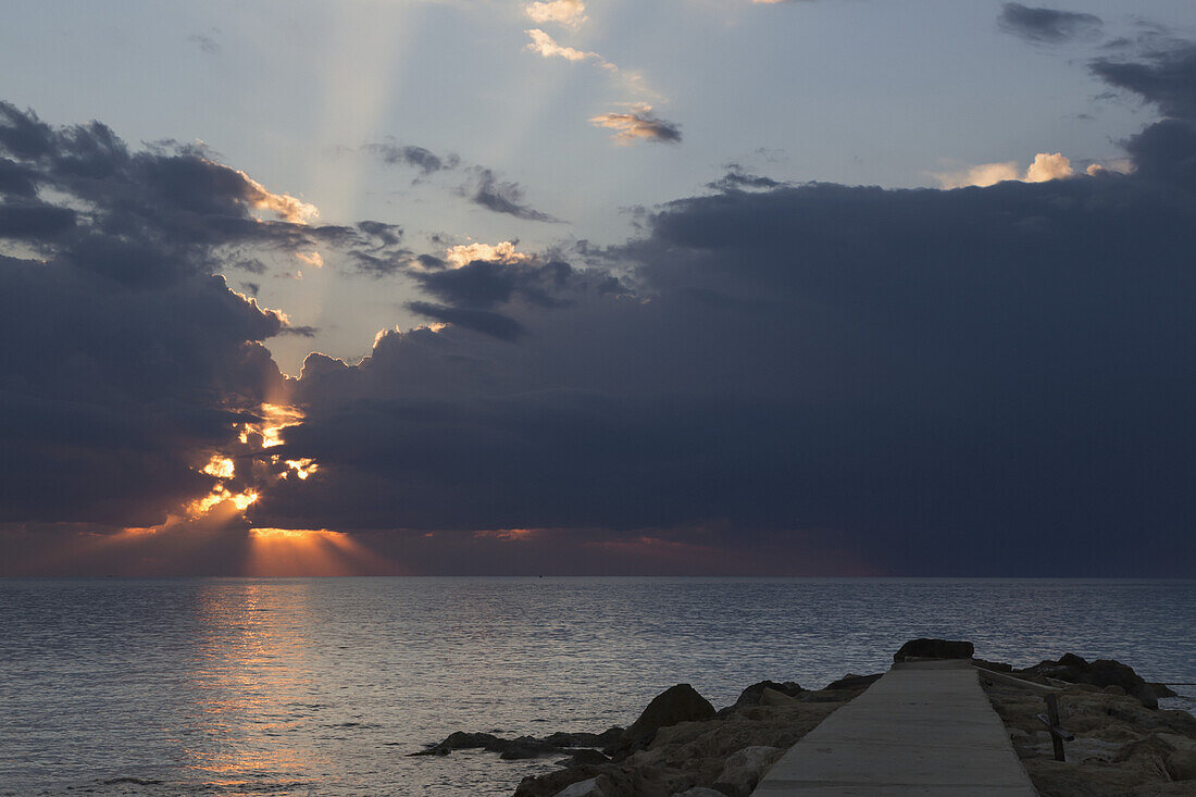 Dramatischer Himmel mit Sonnenstrahlen, die bei Sonnenuntergang über dem Horizont durch dunkle Sturmwolken brechen; Paphos, Zypern
