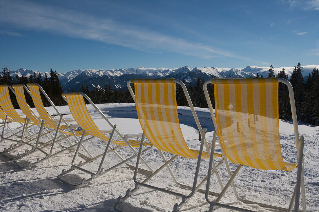 Eine Reihe von gelb-weiß gestreiften Liegestühlen wartet auf Kunden im Sonnenschein vor einem Café am oberen Ende einer Skipiste mit Blick auf die Berggipfel der österreichischen Alpen; Filzmoos, Österreich