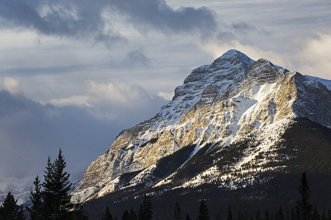 Sun Breaking Through The Clouds Partially Lighting A Snow Covered Mountain With Clouds In The Sky; Kananaskis Country, Alberta, Canada