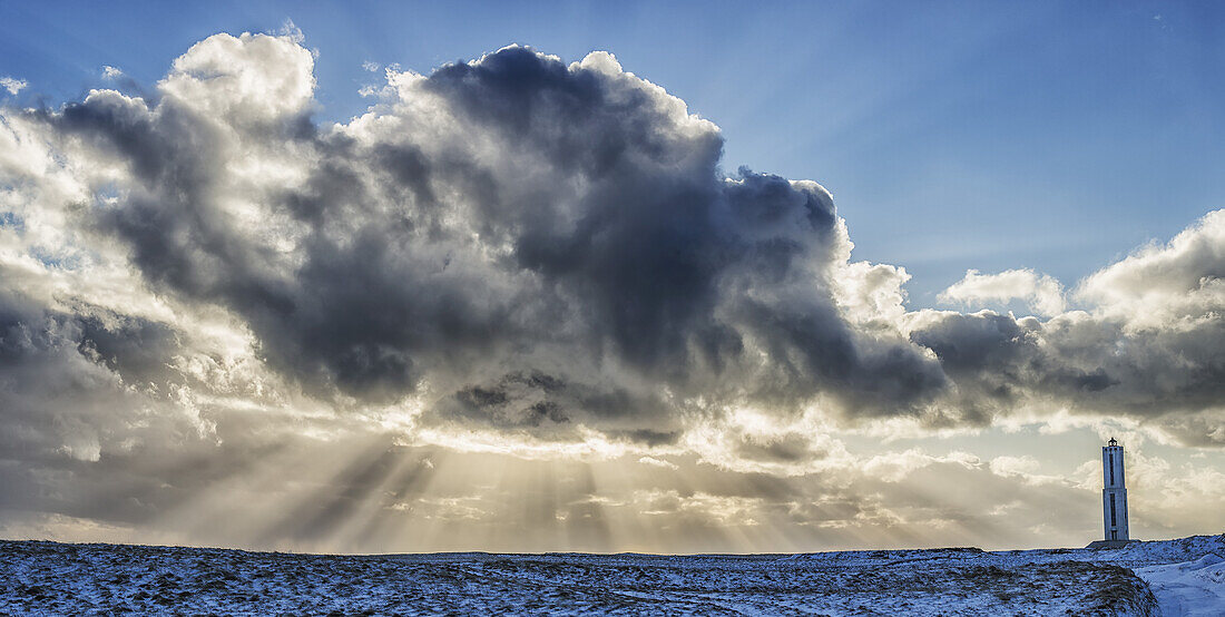 Lichtstrahlen leuchten hinter den Wolken am Himmel über einem Leuchtturm an der Südküste Islands; Island