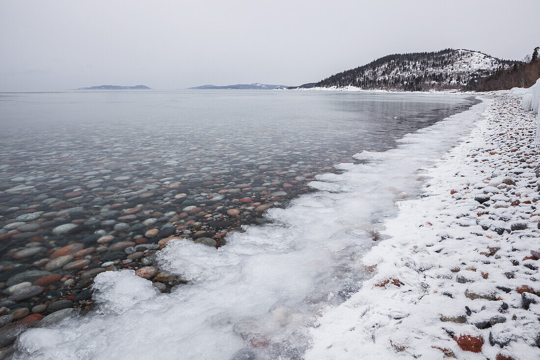 Kieselsteine in ruhigem Wasser mit Eis und Schnee entlang der Küste des Lake Superior